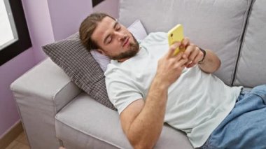 Genç İspanyol adam kanepede uzanıyor ve evde uyumak için akıllı telefon kullanıyor.