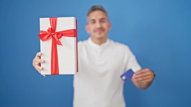 単離された青い背景にクレジットカードと贈り物を保持している若いヒスパニック男性 — ストック動画