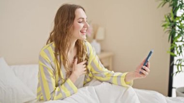 Young blonde woman having video call sitting on bed at bedroom