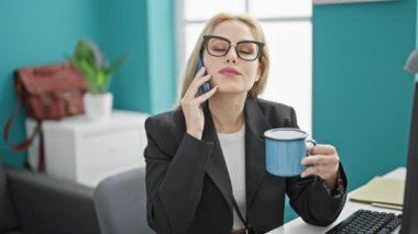 Genç sarışın iş kadını akıllı telefondan konuşuyor ofiste kahve içiyor.