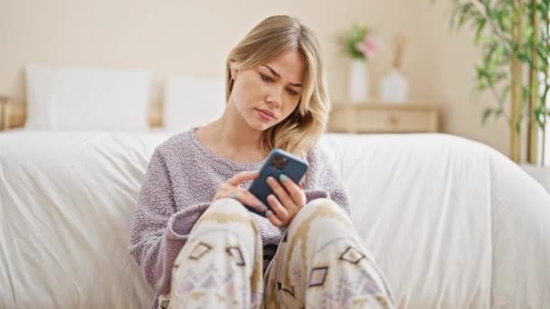 スマートフォンを使用する若い金髪の女性は寝室で床に座っていることを強調しました — ストック動画