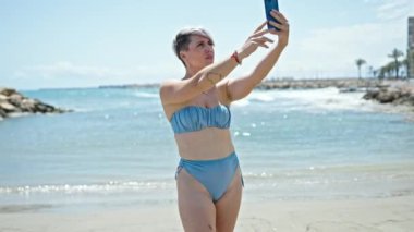 Bikini giyen genç bir turist sahilde akıllı telefondan selfie çekiyor.