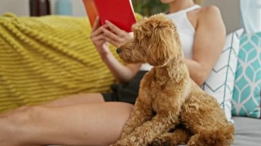 Evde kanepede oturan köpek okuma kitabı olan beyaz bir kadın.