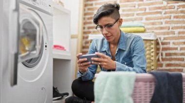 Young beautiful hispanic woman using smartphone waiting for washing machine at laundry room