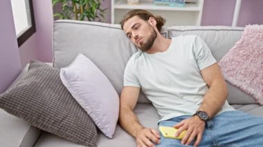 Genç İspanyol adam akıllı telefon kullanıyor. Kanepede oturuyor ve evde üzgün üzgün bakıyor.
