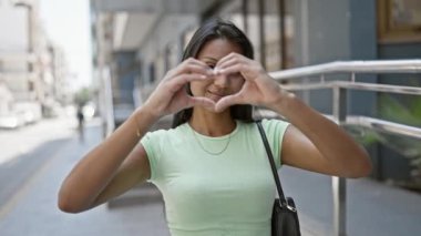 Genç Latin kadın gülümsüyor. Kendine güveniyor. Sokakta elleriyle kalp hareketi yapıyor.