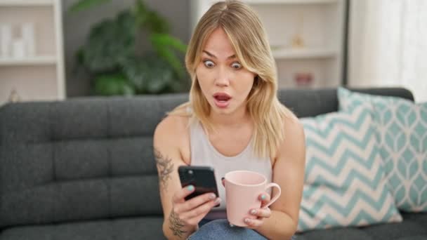 年轻的金发女人在家里用智能手机喝咖啡 脸上带着惊讶的表情 — 图库视频影像