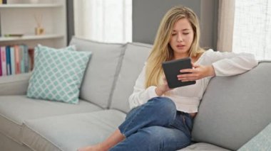 Young blonde woman using touchpad sitting on sofa at home