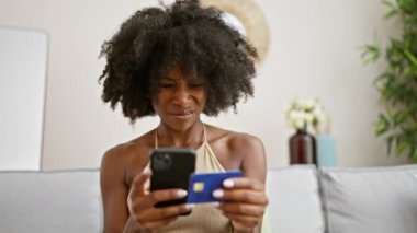 Akıllı telefon ve kredi kartıyla alışveriş yapan Afrikalı Amerikalı kadın evde üzgün görünüyor.