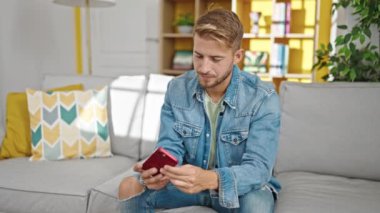 Beyaz bir adam akıllı telefonuyla fotoğraf çekiyor. Evdeki kanepede oturuyor.