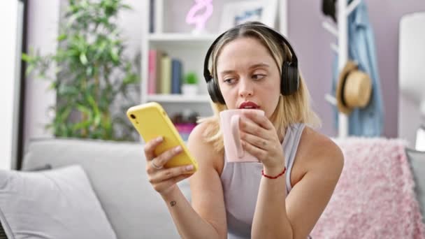 Młoda Blondynka Używa Smartfona Słuchawek Picia Kawy Domu — Wideo stockowe