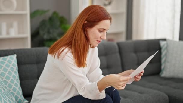 年轻的红头发女人坐在家里的沙发上看书 — 图库视频影像