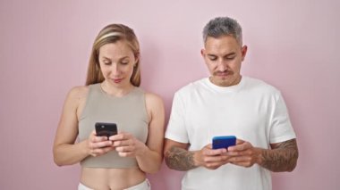 Kadın ve erkek, izole edilmiş pembe arka planda akıllı telefonlar kullanarak yan yana duruyorlar.