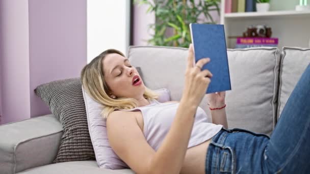 ソファーに横たわる若い金髪の女性は 自宅で寝るために本を読むのをやめます — ストック動画
