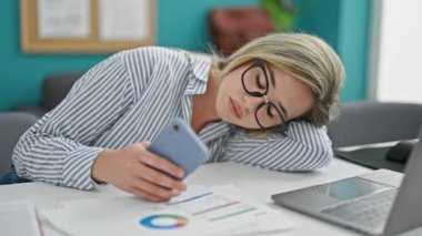 Genç sarışın iş kadını ofiste akıllı telefon kullanarak sıkılmış ve yorgun çalışıyor.
