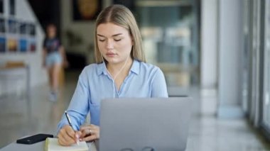 Laptop ve kulaklık kullanan genç sarışın iş kadını ofiste notlar alıyor.