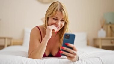 Şehvetli iç çamaşırları giyen genç sarışın kadın yatak odasında akıllı telefon kullanarak yatağa uzanıyor.