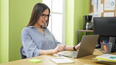 Genç, güzel İspanyol iş kadını. Laptop ve kulaklık kullanıyor. Ofiste birinci sınıf jest yapıyor.