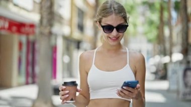 Genç sarışın kadın akıllı telefon kullanıyor. Sokakta kahve içiyor.