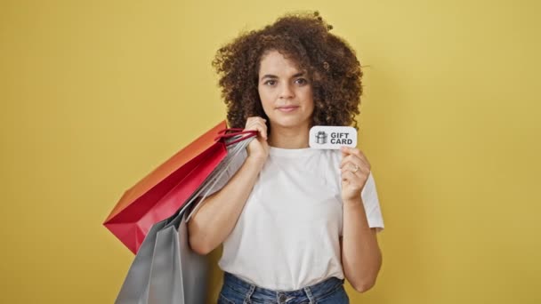 ショッピングバッグとギフトカードを保持している若い美しいヒスパニックの女性は 孤立した黄色の背景に微笑みます — ストック動画