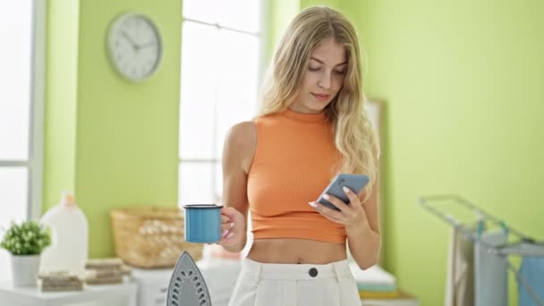 Νεαρή Ξανθιά Γυναίκα Που Χρησιμοποιεί Smartphone Πίνοντας Καφέ Όρθια Σιδερώνοντας — Αρχείο Βίντεο