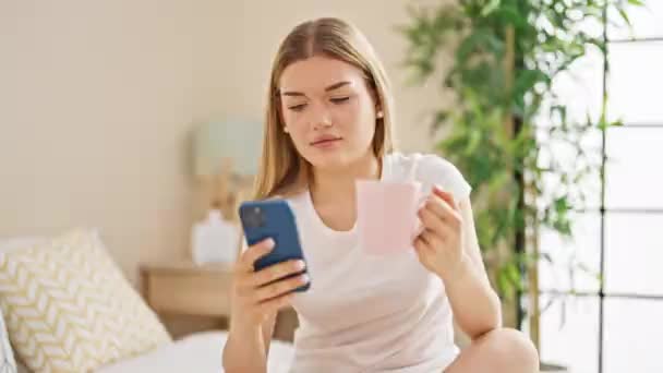 若いですブロンド女性使用スマートフォン飲料コーヒーでベッドルーム — ストック動画