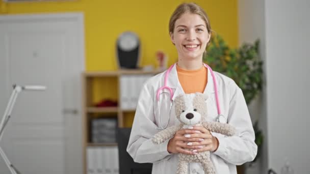 Молода Блондинка Лікар Посміхається Впевнено Тримає Плюшевого Ведмедя Клініці — стокове відео