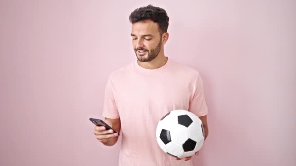 Jovem Hispânico Usando Smartphone Segurando Bola Futebol Sobre Fundo Rosa — Vídeo de Stock