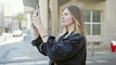 Sokakta gülümseyen akıllı telefonun yanında video kaydeden genç sarışın kadın.