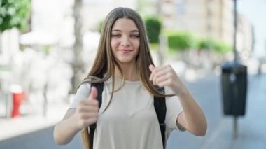 Sırt çantası takan genç ve güzel bir kız sokakta baş parmak hareketi yapıyor.