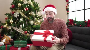 Yılbaşı şapkası takan genç kel bir adam evde şaşırmış yüzüyle hediyesini açıyor.