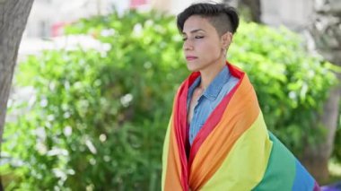 Parkta gökkuşağı bayrağı takmış, ciddi bir ifadeyle duran genç, güzel İspanyol kadın.