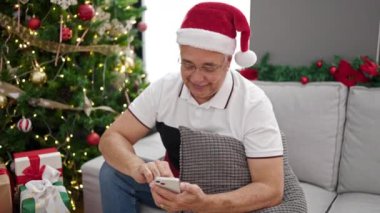 Beyaz saçlı, akıllı telefon kullanan orta yaşlı bir adam evdeki Noel ağacının yanında koltukta oturuyor.