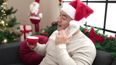 Genç Çinli bir adam evde Noel ağacının yanında akıllı telefondan kahve içiyor.