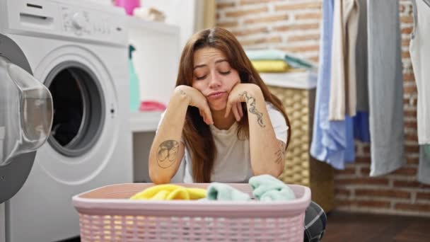 若い美しいヒスパニックの女性は 洗濯室で悲しい表情で服でバスケットに乗っています — ストック動画