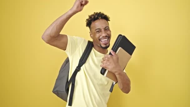 隔離された黄色の背景を祝う本を保持するバックパックを身に着けているアフリカ系アメリカ人男性の学生 — ストック動画