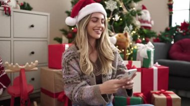 Young blonde woman using smartphone sitting by christmas tree at home