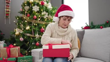 Olgun İspanyol kadın Noel şapkası takıyor, hediyesini açıyor ve evine üzgün bakıyor.