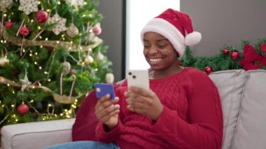 Afrikalı Amerikalı bir kadın evde akıllı telefonuyla Noel alışverişi yapıyor.