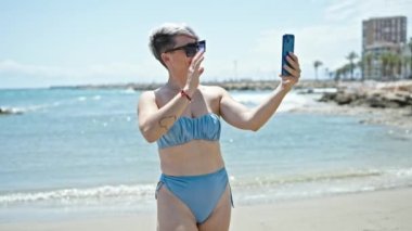 Bikini giyen ve güneş gözlüğü takan genç bir turist kumsalda video görüşmesi yapıyor.