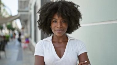 Afro-Amerikalı kadın gülümsüyor. Kollarını kavuşturup caddede duruyor.