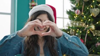 Genç ve güzel İspanyol kadın Noel ağacının yanında gülümsüyor. Elleriyle kalp şekli yapıyor.