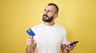 Akıllı telefon ve kredi kartıyla alışveriş yapan İspanyol genç adam izole edilmiş sarı arka plan üzerinde fikir yürütüyor.
