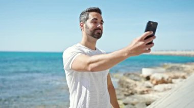 Genç İspanyol gülümseyen adam sahilde akıllı telefondan özçekim yapıyor.