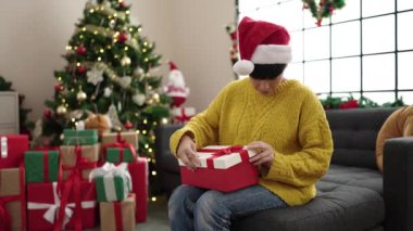 Genç Çinli bir kadın hediye paketini açıyor evde Noel ağacının yanında oturuyor.