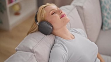 Evdeki kanepede rahatça müzik dinleyen genç sarışın kadın.