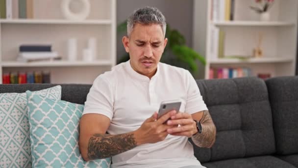 Ung Latinamerikan Man Med Smartphone Ser Upprörd Hemma — Stockvideo