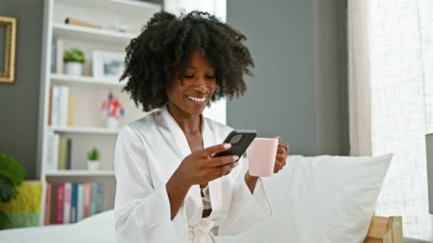 アフリカ系アメリカ人女性がベッドに座って寝室でスマートフォンを使用してコーヒーを飲む — ストック動画