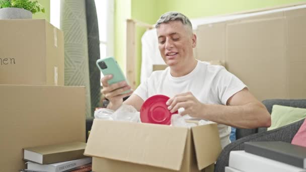 Jovem Caucasiano Homem Desembalagem Caixa Papelão Usando Smartphone Nova Casa — Vídeo de Stock