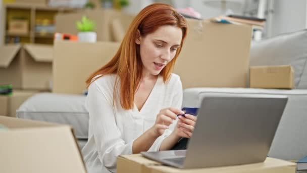 若い赤毛の女性は 新しい家で床に座ってラップトップとクレジットカードで買い物 — ストック動画